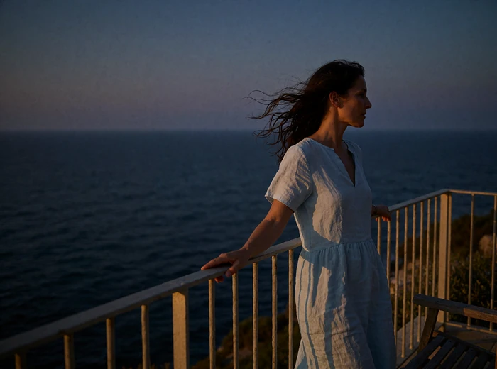 Attractive woman in a pale blue linen dress standing at the railing of a coastal terrace at dusk