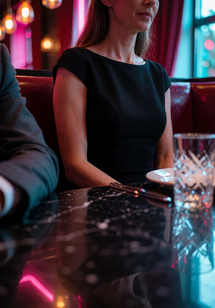 Woman in a black cocktail dress seated at a dark private dining booth opposite a man in a tailored suit